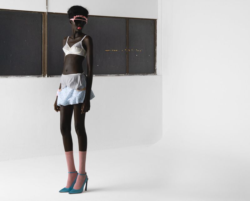 Full-length studio shot of a Black woman in lingerie, pink socks, and heels, standing against a stark white background and window.