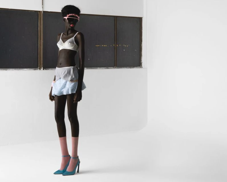 Full-length studio shot of a Black woman in lingerie, pink socks, and heels, standing against a stark white background and window.