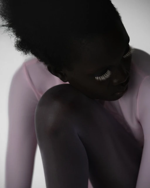 Close-up of a dark-skinned adult female with long, white eyelashes, wearing a pink, long-sleeved top, captured in a photo studio.