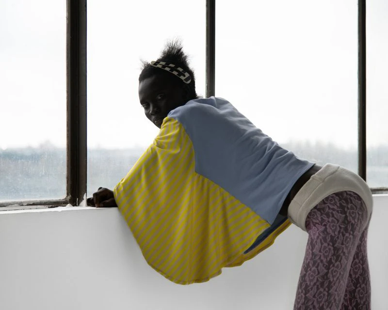 Dark-skinned woman in colorful clothing leans by window with natural light. She wears patterned leggings, a top with yellow sleeves and blue panel.