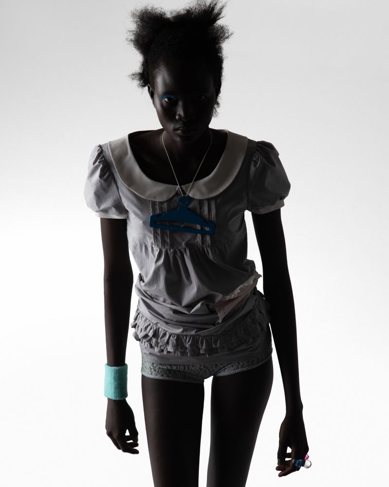 Silhouette of a Black female adult fashion model in a grey baby-doll style outfit with a blue necklace against a white background.