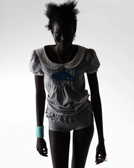 Silhouette of a Black female adult fashion model in a grey baby-doll style outfit with a blue necklace against a white background.