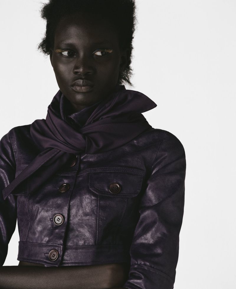 Fashion portrait of a Black female adult in a short purple leather jacket, posed against a white background in eye-level studio light.