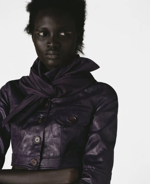 Fashion portrait of a Black female adult in a short purple leather jacket, posed against a white background in eye-level studio light.