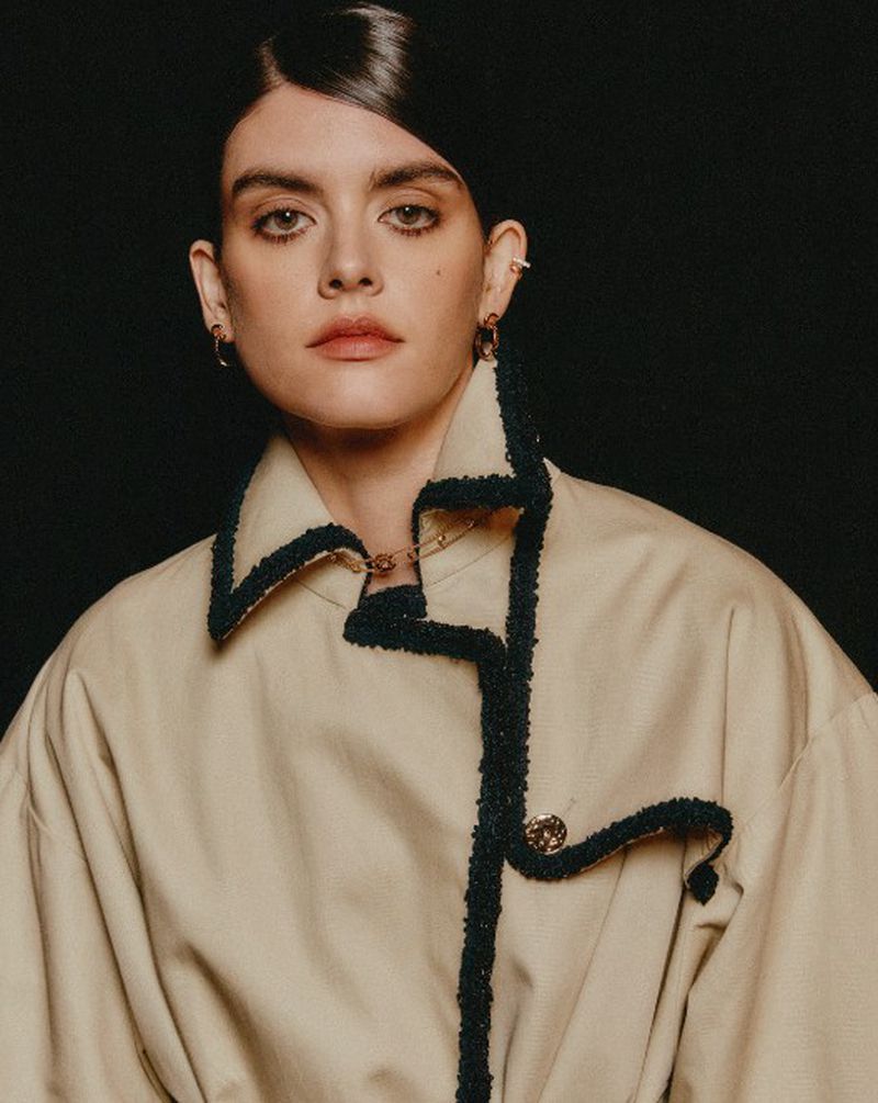 Close-up portrait of a young adult woman with dark hair styled to the side, wearing a tan jacket with dark trim against a black background.