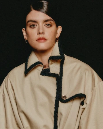 Close-up portrait of a young adult woman with dark hair styled to the side, wearing a tan jacket with dark trim against a black background.