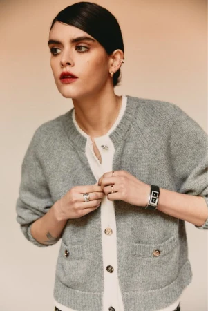 A young, adult woman is shown from the waist up, buttoning a grey knit cardigan, with a neutral colored background, and wearing a watch.