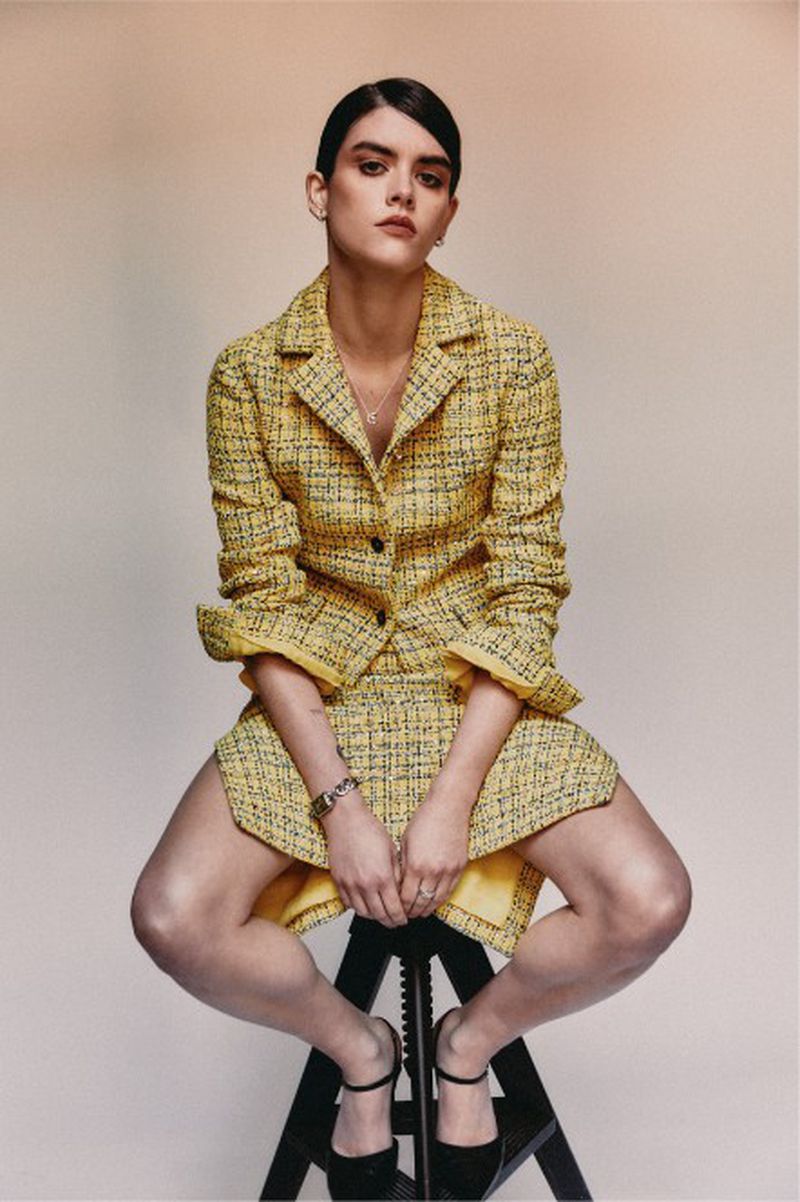 Young woman in a yellow tweed jacket and skirt sits on a black stool against a neutral background in a studio fashion shot.