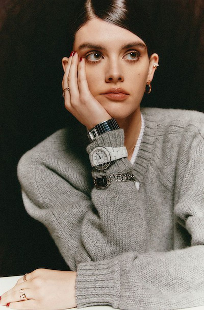 Attractive female model with hand on face, wearing a gray sweater, multiple wristwatches, rings, and earrings against a dark background.