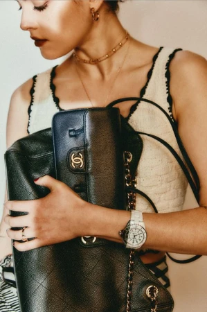 Woman in a cropped top and jewelry holds two black Chanel handbags and shows a white watch against a bright neutral background.