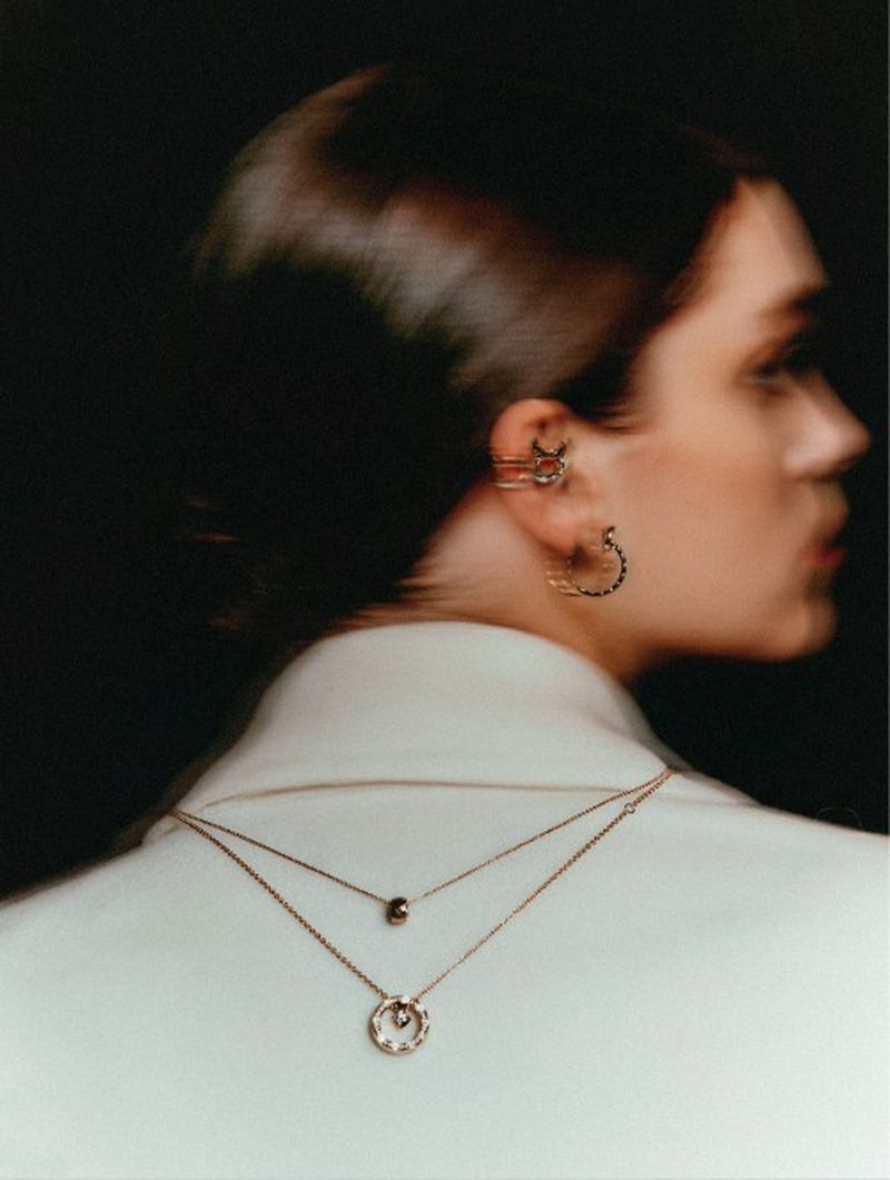 Woman in profile with ear cuff and layered necklaces, wearing a white jacket against a dark background, beauty shot.