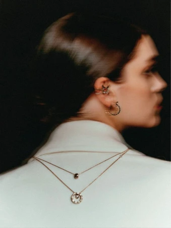 Woman in profile with ear cuff and layered necklaces, wearing a white jacket against a dark background, beauty shot.