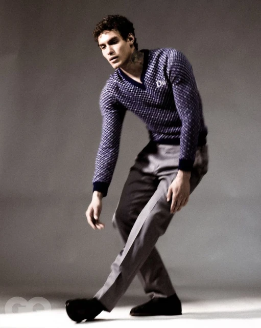 Male fashion model poses in a patterned sweater and gray trousers in a studio setting, captured in a fashion editorial style.