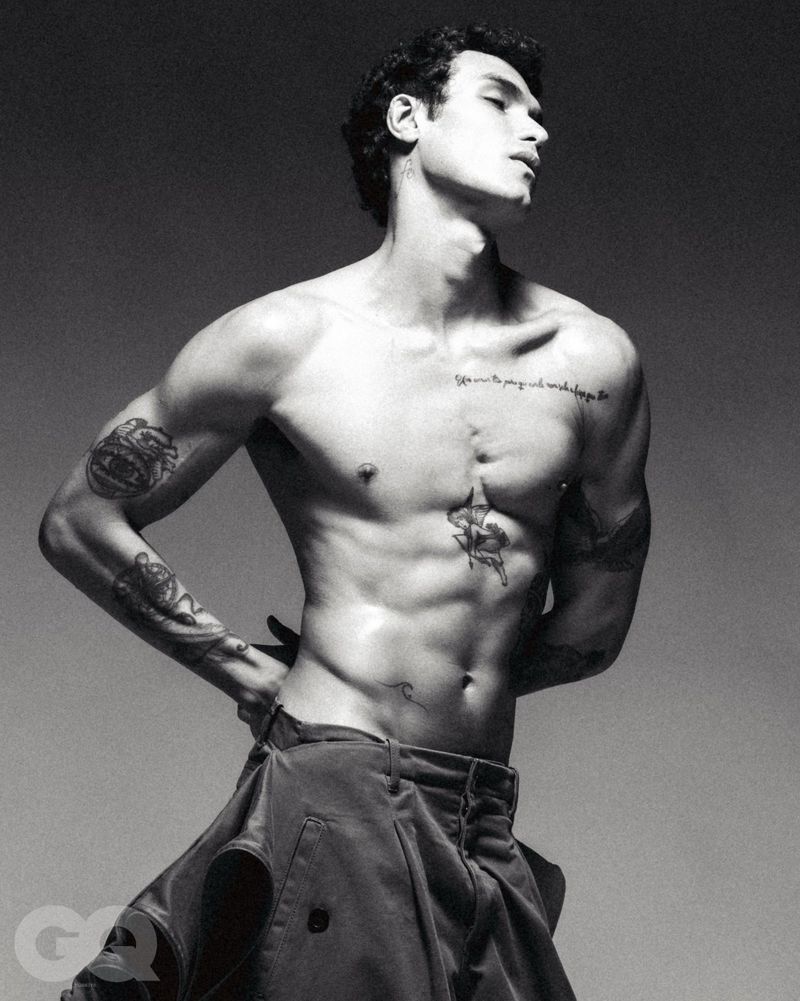 Black and white portrait of a shirtless adult male with tattoos posing in a studio wearing trousers with hands on waist.