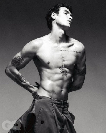 Black and white portrait of a shirtless adult male with tattoos posing in a studio wearing trousers with hands on waist.