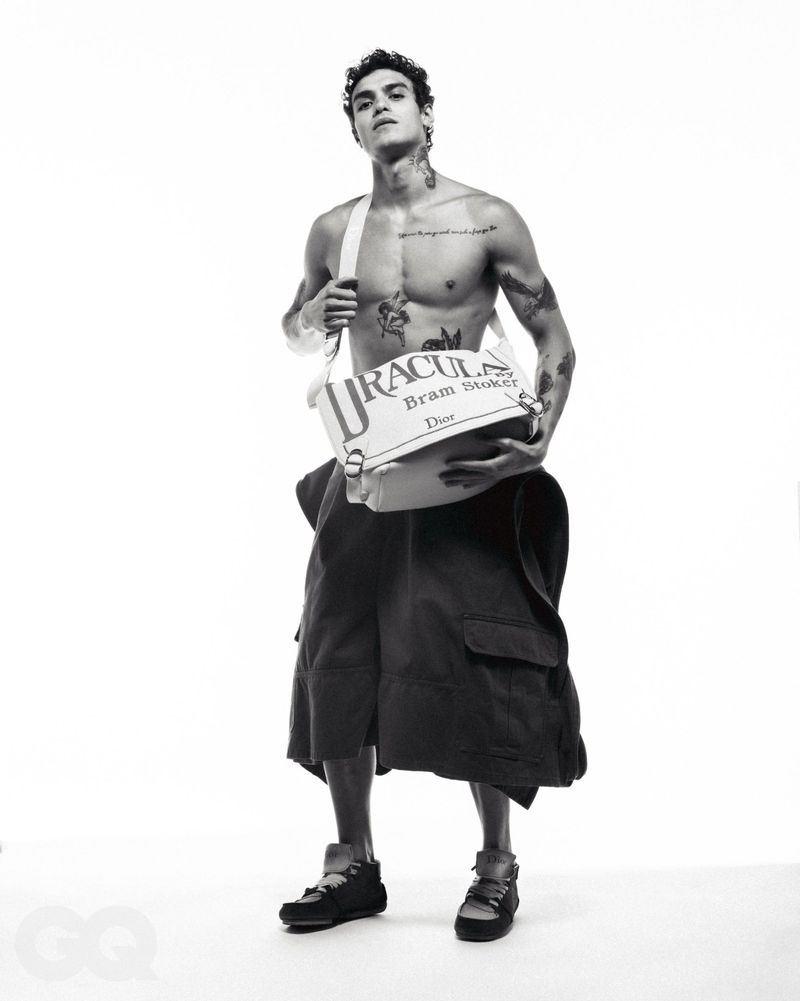 Shirtless man in wide black shorts holds a white Dracula-themed bag, posing in a black-and-white studio portrait on a white backdrop.