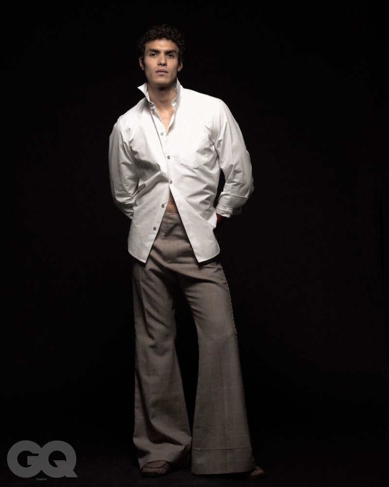 Portrait of a male model in a white shirt and grey flared trousers standing against a black background, lit with studio lighting.