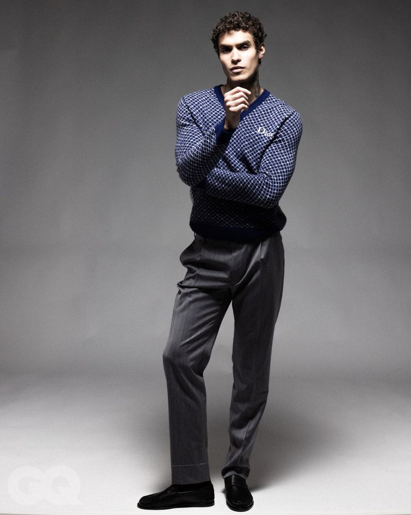 Full shot of a dark-haired adult male fashion model in gray trousers and a blue sweater in a studio with a neutral gray backdrop.