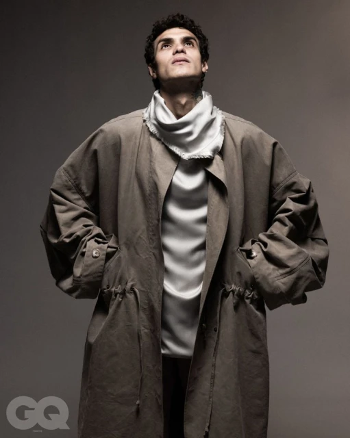 Adult male in earth-tone trench coat and white scarf looks upward in a studio portrait with soft lighting and a neutral backdrop.