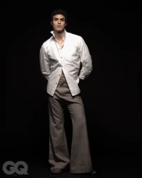 Full-length portrait of a male model wearing an unbuttoned white shirt and grey trousers against a black background.