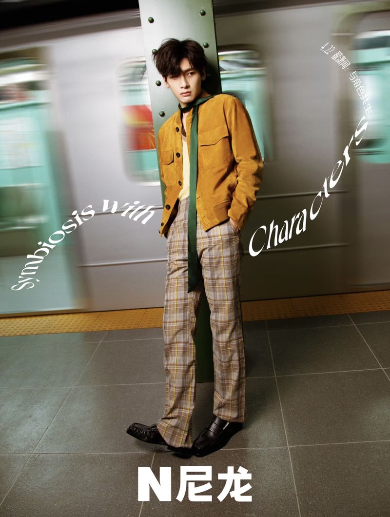 Stylish man in corduroy jacket and plaid pants poses against a blurred train car and subway platform background in a fashion shot.