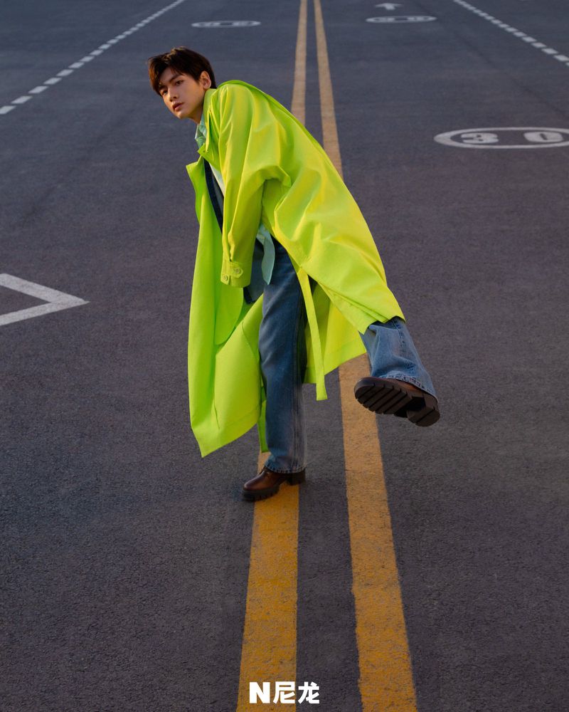 Stylish young man in a neon yellow raincoat and blue jeans poses on an asphalt road marked with bright yellow lines in an urban setting.