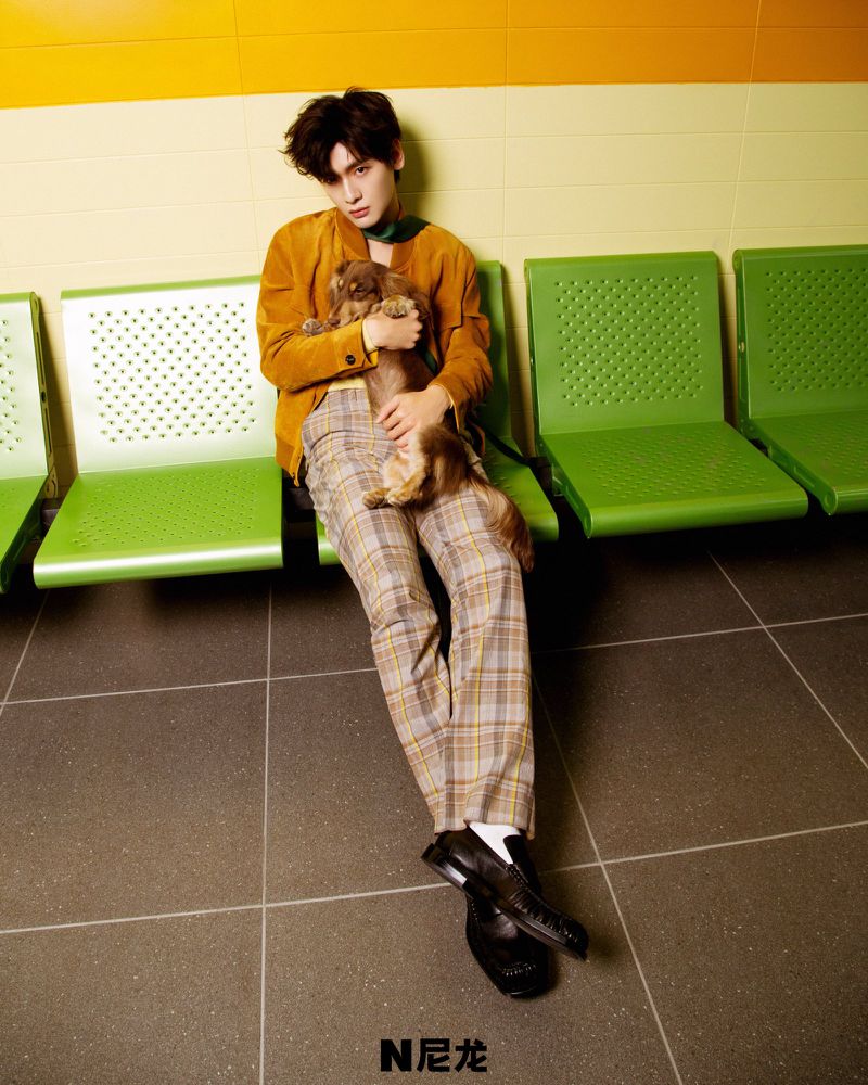 Stylish young man in corduroy jacket poses indoors with his long-haired dachshund. Model seated on bright green indoor public seating.