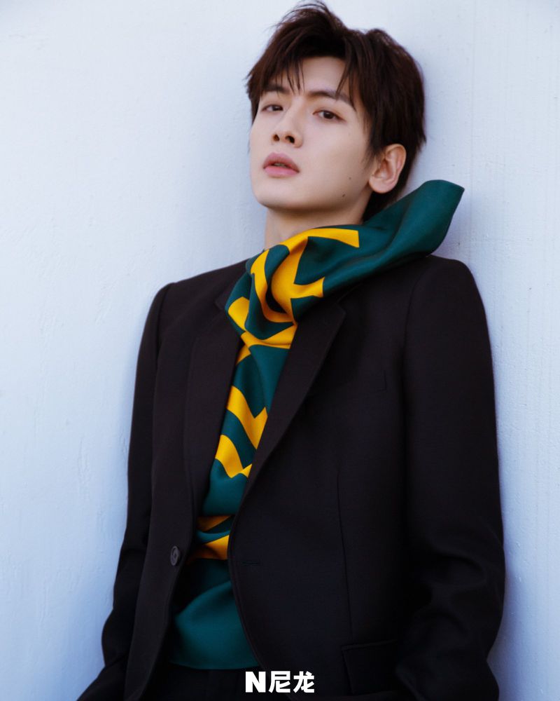 Portrait of a male fashion model in a black suit with a green and yellow patterned scarf, posing against a light blue wall in a studio.