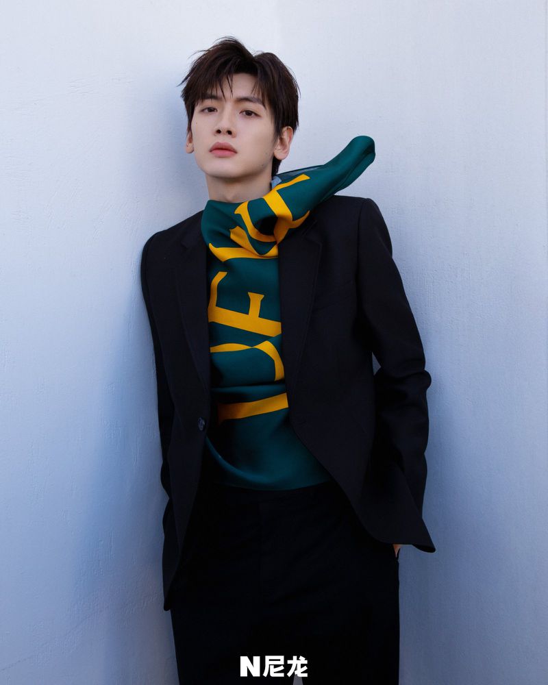 Portrait of a young adult male wearing a black suit with a green and yellow scarf, posing against a light blue wall in a studio setting.