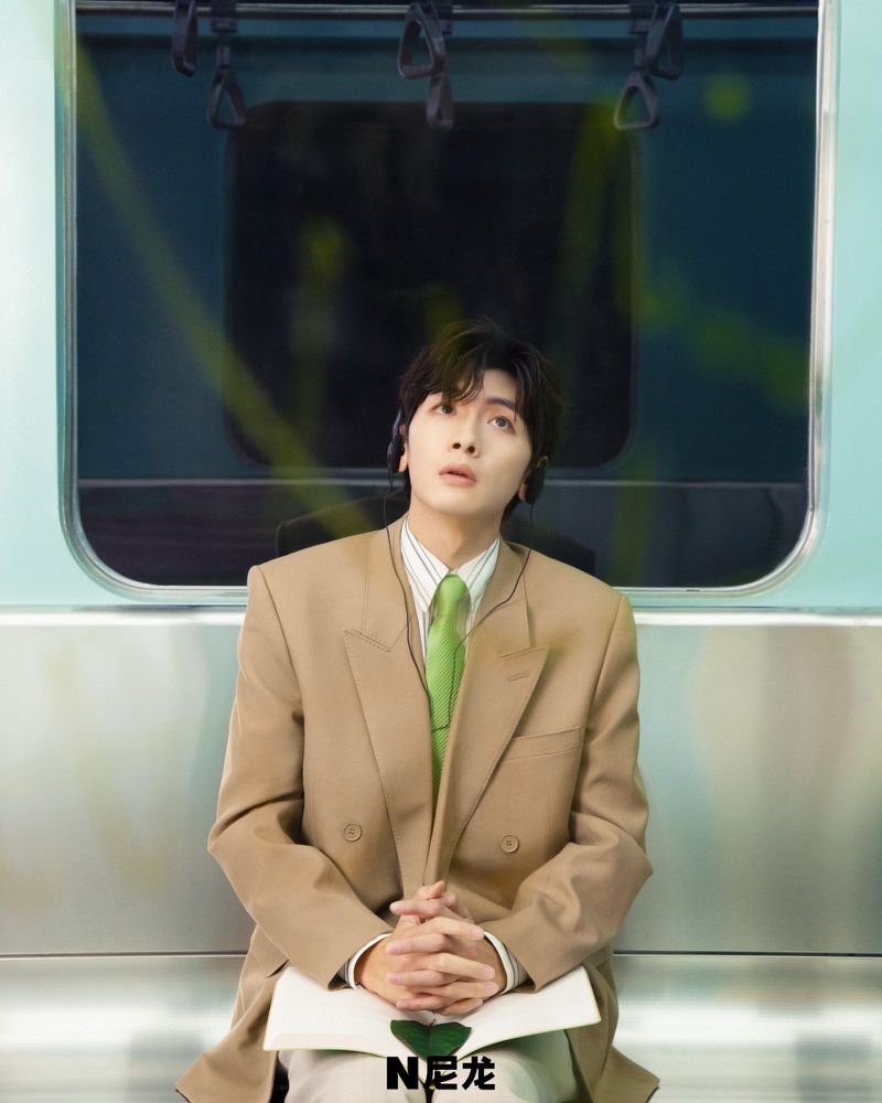 Adult male in beige suit sits in a light blue train car with silver walls, wearing headphones, looking up with neutral expression, holding notepad.