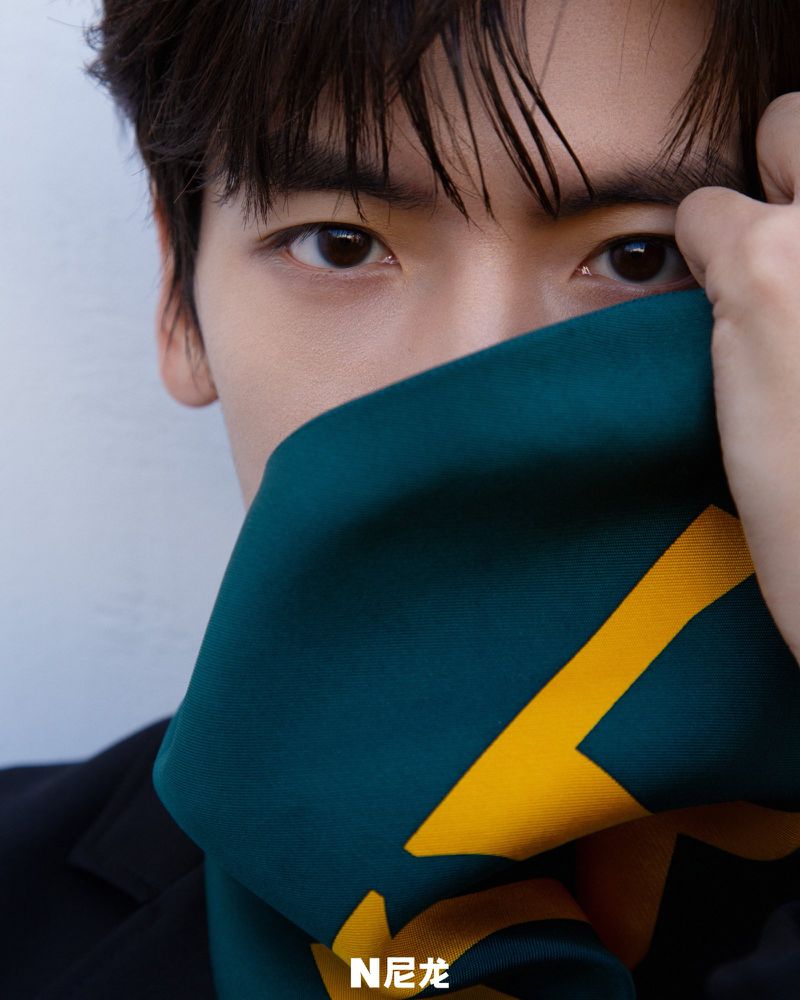 Close-up portrait of a young adult man with dark hair, wearing a dark jacket and a green patterned scarf held to his face.