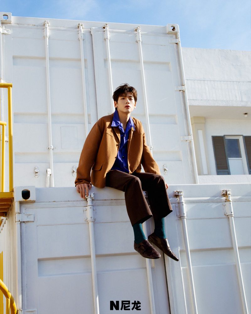 A young Asian man in a brown jacket and brown pants sits atop a white container in an outdoor setting under a blue sky.