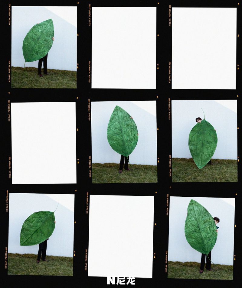 A person stands on grass with a large green leaf in front of a white wall, showcasing fashion and nature in a unique display.