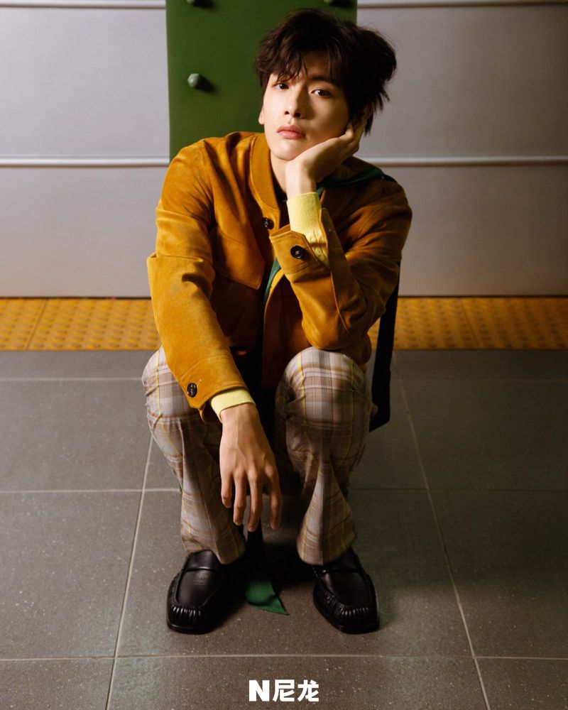 Adult male crouching indoors, wearing an amber jacket and plaid pants, with a muted green and grey background.