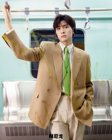An Asian man in a beige suit stands in a subway car, holding onto a rail. He wears a green tie and has black earphones on.