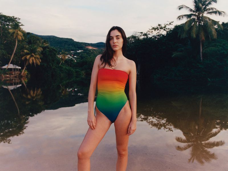 Adult woman in rainbow-colored bathing suit stands by water, with reflection, against tropical background with trees and sky.