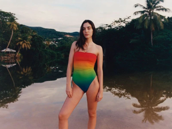 Adult woman in rainbow-colored bathing suit stands by water, with reflection, against tropical background with trees and sky.