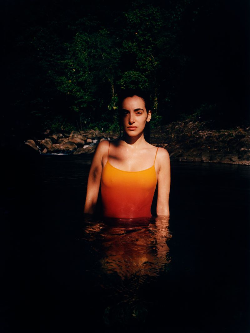 Woman in ombre swimsuit stands in dark water, framed against a forest backdrop, sunlight highlighting the face and upper body.