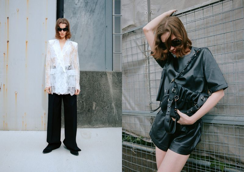 Two fashion models pose outdoors. One wears a white lace jacket with black pants, while the other dons a black leather outfit with a bag.