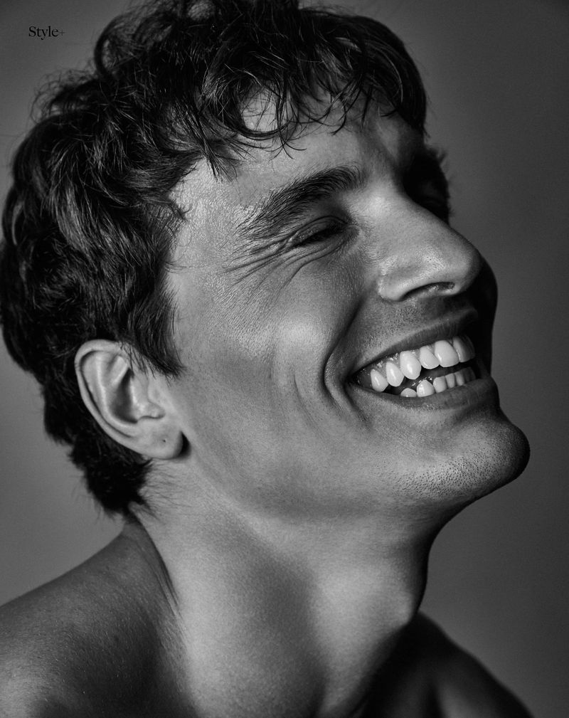 Monochrome close-up portrait of a smiling young adult man with curly dark hair, angled facial pose, and neutral grey background.