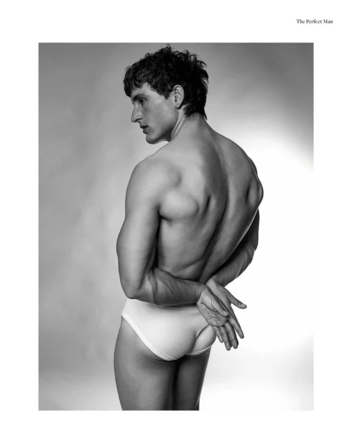 Black and white portrait of a shirtless man in briefs, facing left with hands clasped behind his back, against a soft, gradient background.