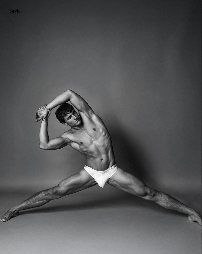 Striking black and white studio shot of a muscular adult male doing a side stretch, torso exposed, posing against a gray backdrop.