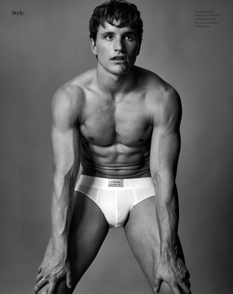 Black and white studio portrait of a muscular male model with an athletic build, wearing briefs, kneeling with hands on knees.