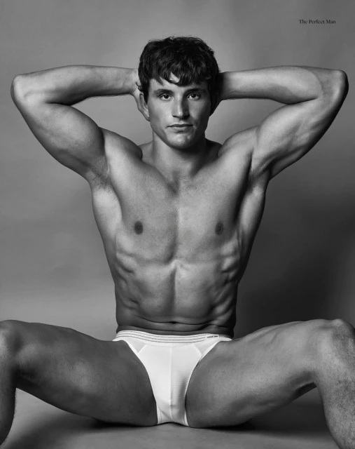 Striking black and white studio portrait of a muscular adult male, shirtless, wearing briefs, and seated against a gray backdrop.