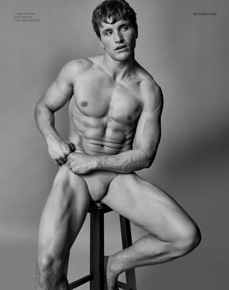 Striking black and white studio portrait of a muscular adult man posing on a stool, showcasing fitness and fashion in an editorial style.
