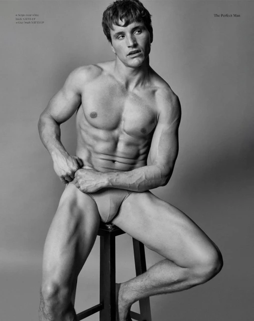 Striking black and white studio portrait of a muscular adult man posing on a stool, showcasing fitness and fashion in an editorial style.