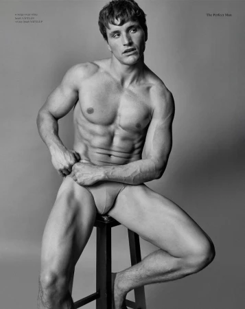 Striking black and white studio portrait of a muscular adult man posing on a stool, showcasing fitness and fashion in an editorial style.