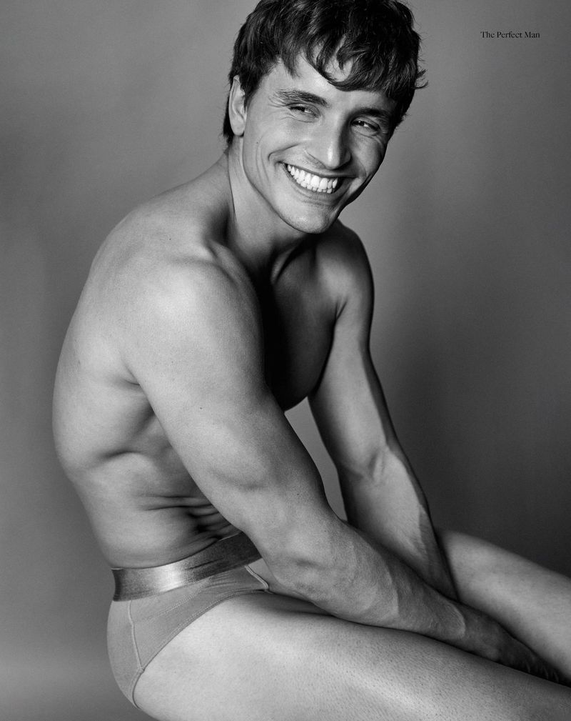 Monochrome portrait of a smiling, muscular adult male with dark hair sitting against a solid gray background in minimal clothing.