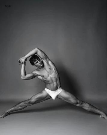 Black and white studio portrait of male model Sergio Modrego stretching in white underwear against a grey background.