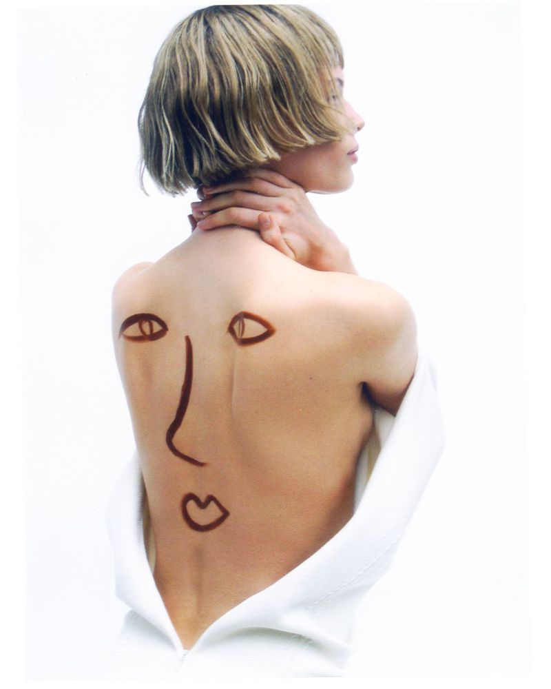 Woman with short blonde hair faces away, a drawn face on her bare back, wearing a white open-back shirt, against a white background.